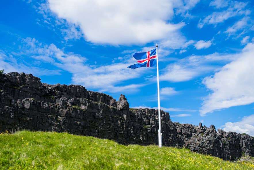 Icelandic flag stands near the rocky cliffs of Thingvellir National Park, marking the rift between tectonic plates under a blue sky.