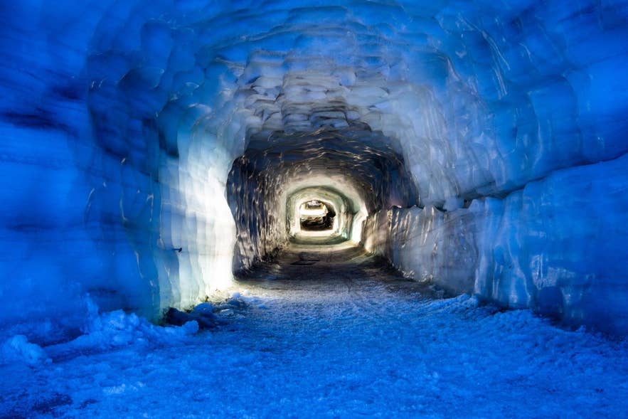 A brightly lit man-made ice tunnel inside Langjokull Glacier, with smooth blue walls and a snowy floor leading toward the tunnel’s illuminated exit.