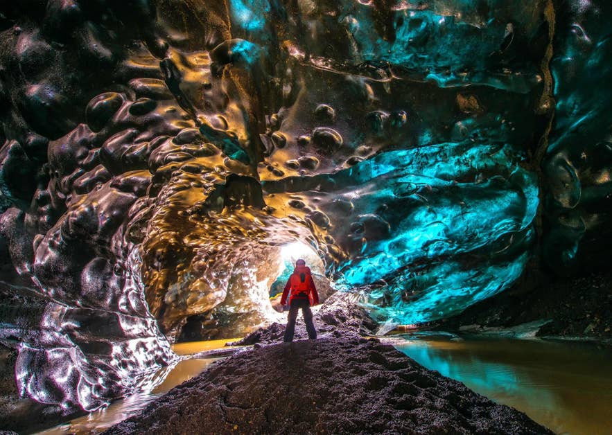 Visitor in a red jacket silhouetted against daylight at the end of a golden and turquoise ice cave tunnel in Iceland.