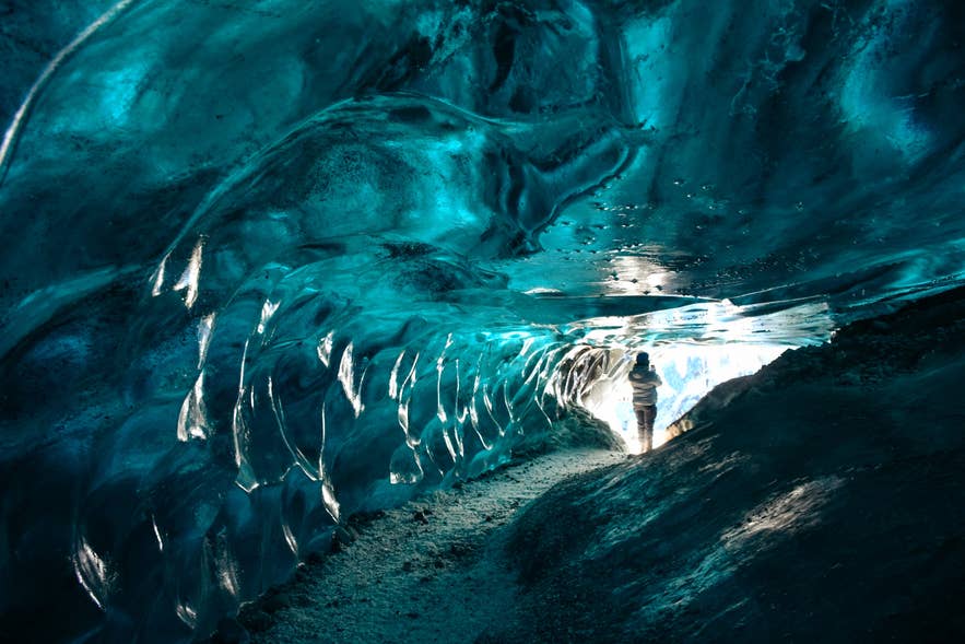 Person walking through a glowing blue ice cave in Iceland with smooth glacier walls and natural light at the entrance.