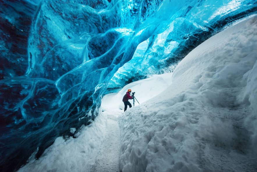 A photographer inside a blue ice cave in Iceland, setting up a camera on deep snow beneath the cave’s glowing, sculpted ice ceiling.