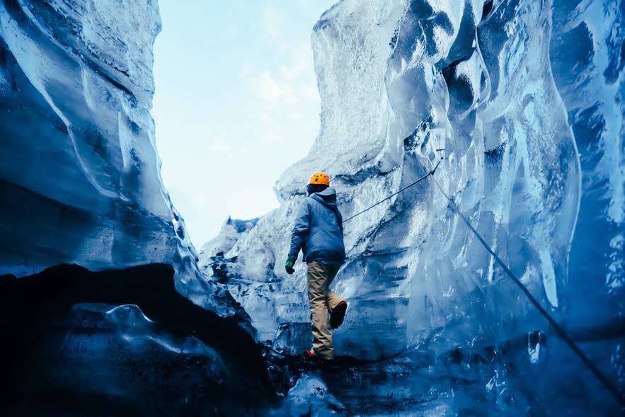 Guide in a helmet climbing a rope-lined path between towering walls of glacier ice in Iceland.