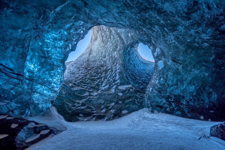 Twin arched openings letting daylight into a blue ice cave in Iceland, with snow on the cave floor.