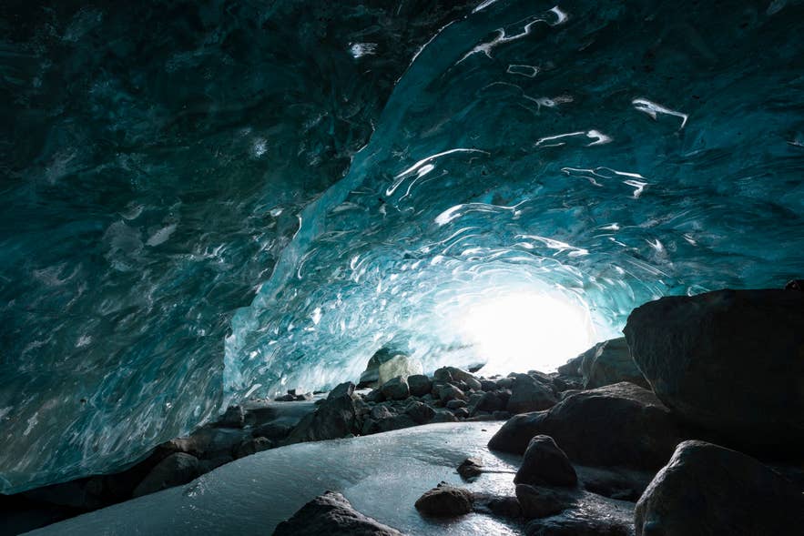 Daylight flooding through the entrance of a deep blue ice cave in Iceland onto a rocky floor.