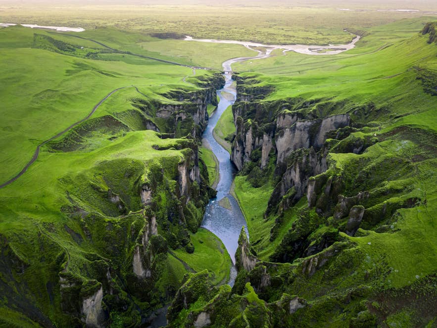 Aerial view of a deep mossy canyon in Iceland with a winding river, steep cliffs, and bright green surrounding hills.