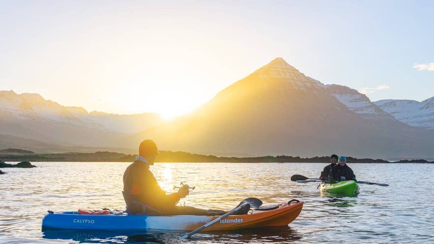 Persona che pratica kayak sulle acque calme islandesi in agosto, con montagne innevate illuminate dal sole della sera estiva.