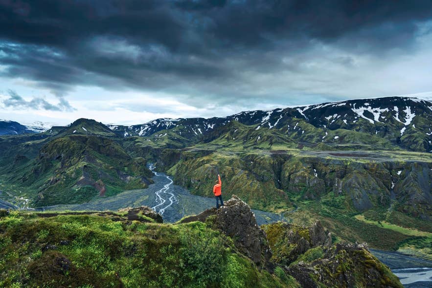 A hiker in a red jacket overlooks Iceland’s dramatic green mountains and glacier rivers under a moody summer sky.