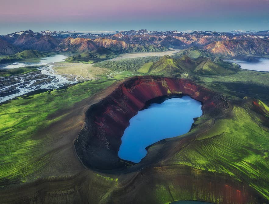 Aerial view of a volcanic crater lake in Iceland, surrounded by green moss, red slopes, braided rivers, and distant mountains.