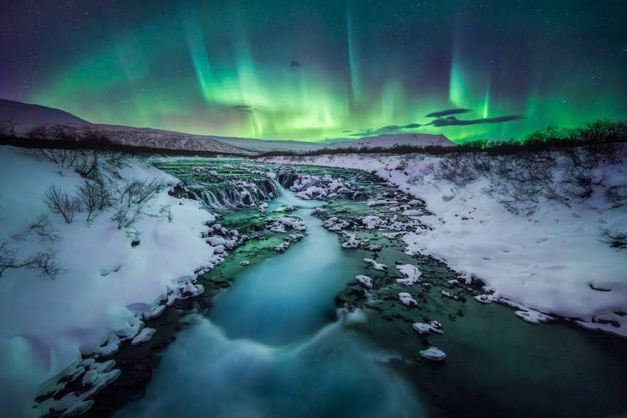 Northern lights dance above a snowy Icelandic river and frozen waterfall, glowing green across the winter night sky.