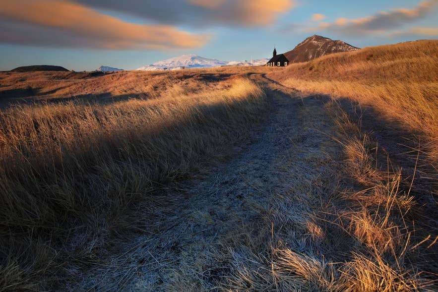 Golden hour light hits a winding path to the black Budir Church in Iceland, with snowy mountains in the distance.
