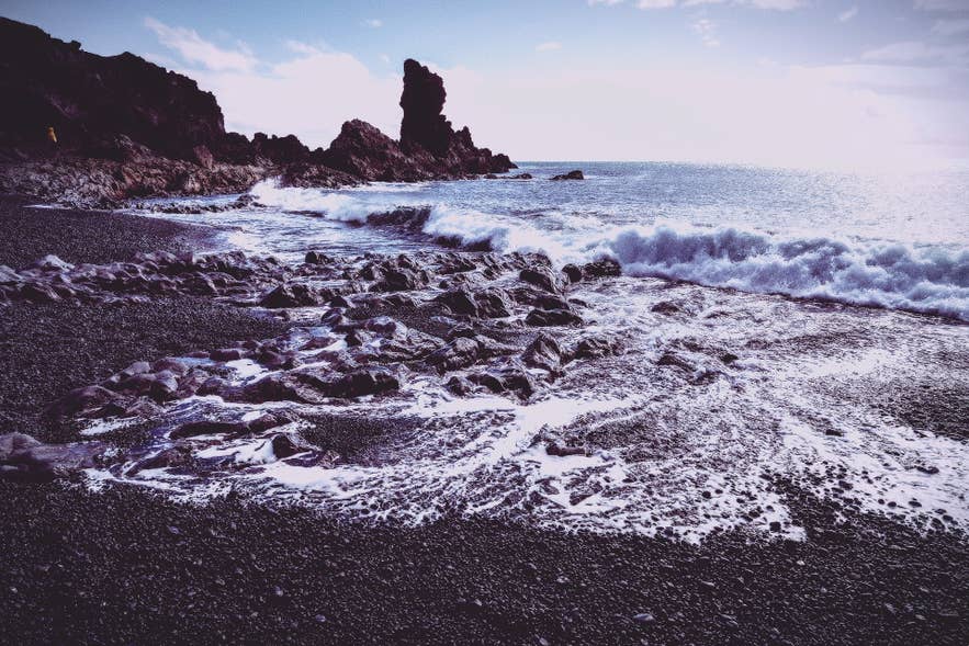 Waves wash over black pebbles and lava rocks on Djupalonssandur Beach with jagged cliffs rising in the background.