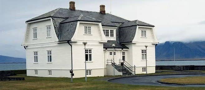 The white wooden Hofdi House stands by the sea with a gray roof and mountains in the background.