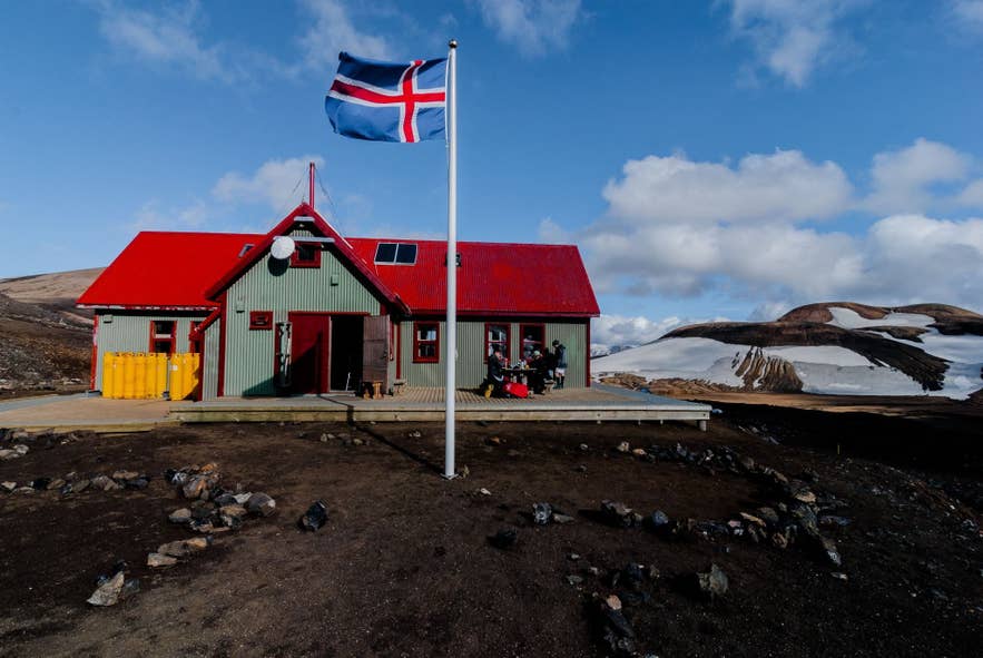 Icelandic flag waves in front of a red-roofed mountain hut with snowy hills and rocky ground under a bright blue sky.