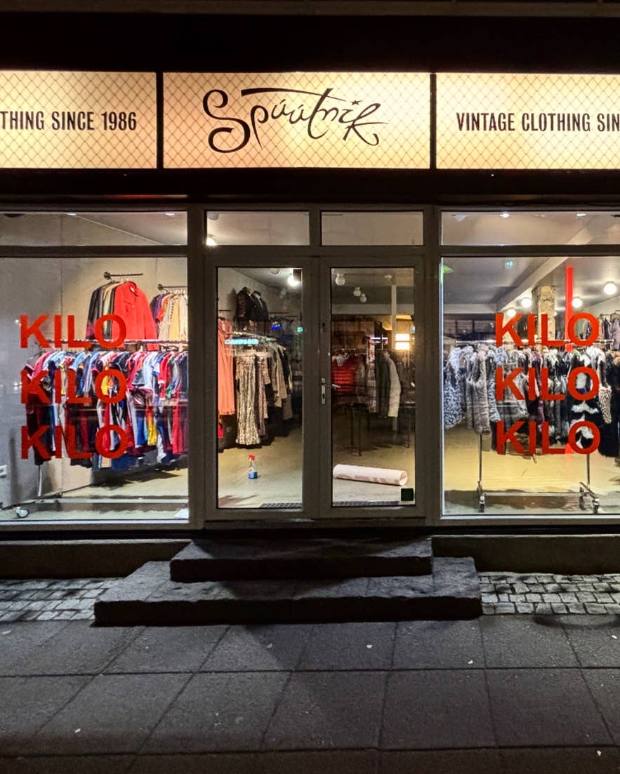 Front entrance of Spúútnik vintage clothing store in Reykjavik with illuminated signage and racks of secondhand fashion visible through the windows at night.