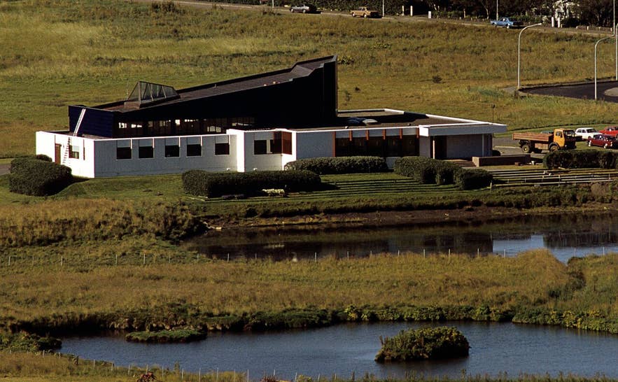 Modern Nordic House in Reykjavík with white sections, angular dark roof, pond reflection, and green lawns under bright sky.