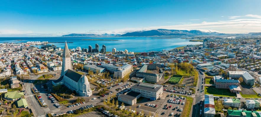 Aerial view of Reykjavík with Hallgrímskirkja’s tall spire, colorful cityscape, coastline, and snowy mountains.