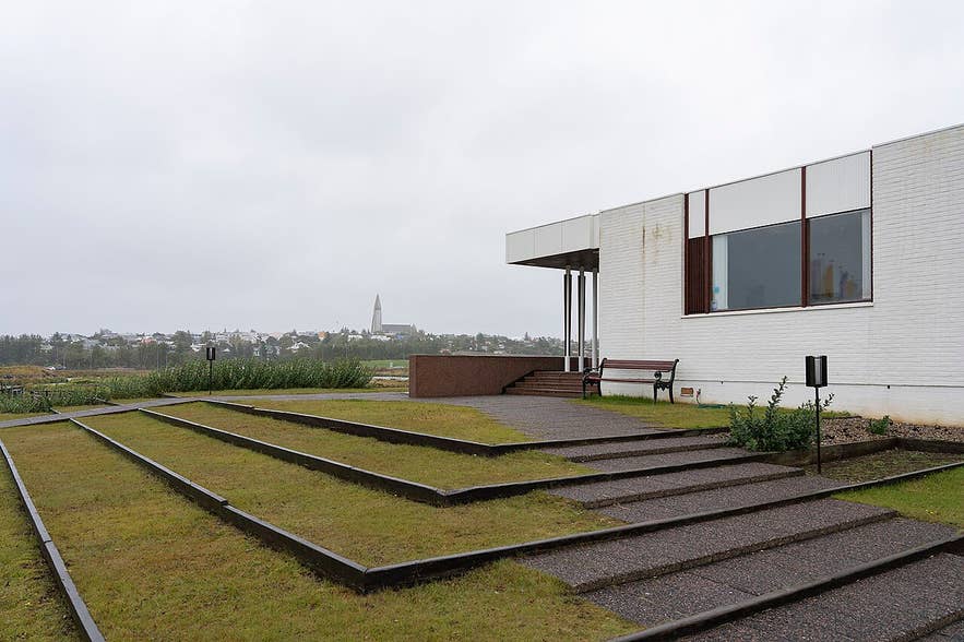 Nordic House in Reykjavík with minimalist white brick walls, tiered grassy platforms, and view of town skyline.