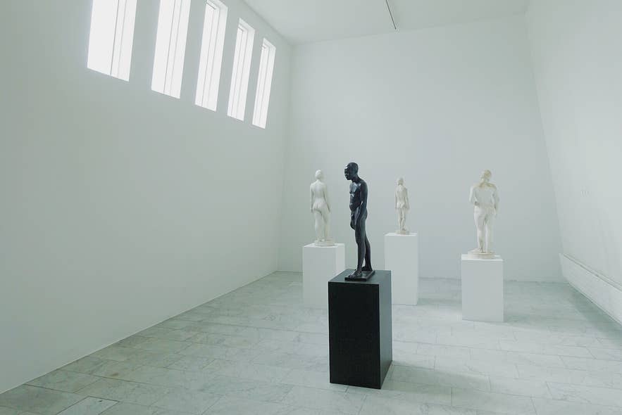 Black statue displayed on a pedestal in a bright white gallery with classical sculptures and tall windows.
