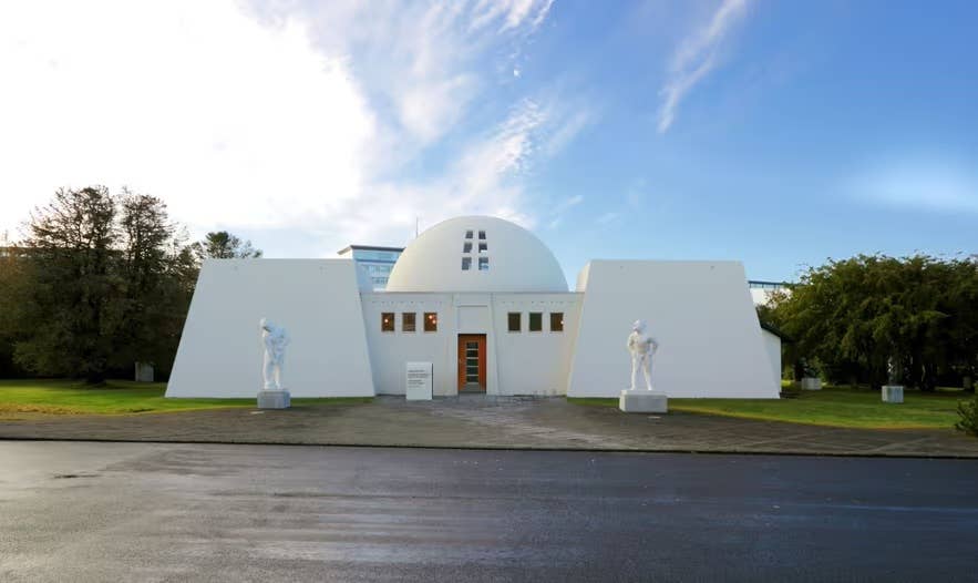 White Ásmundarsafn museum building in Reykjavik, with two sculptures outside, surrounded by green lawns and trees under a blue sky.