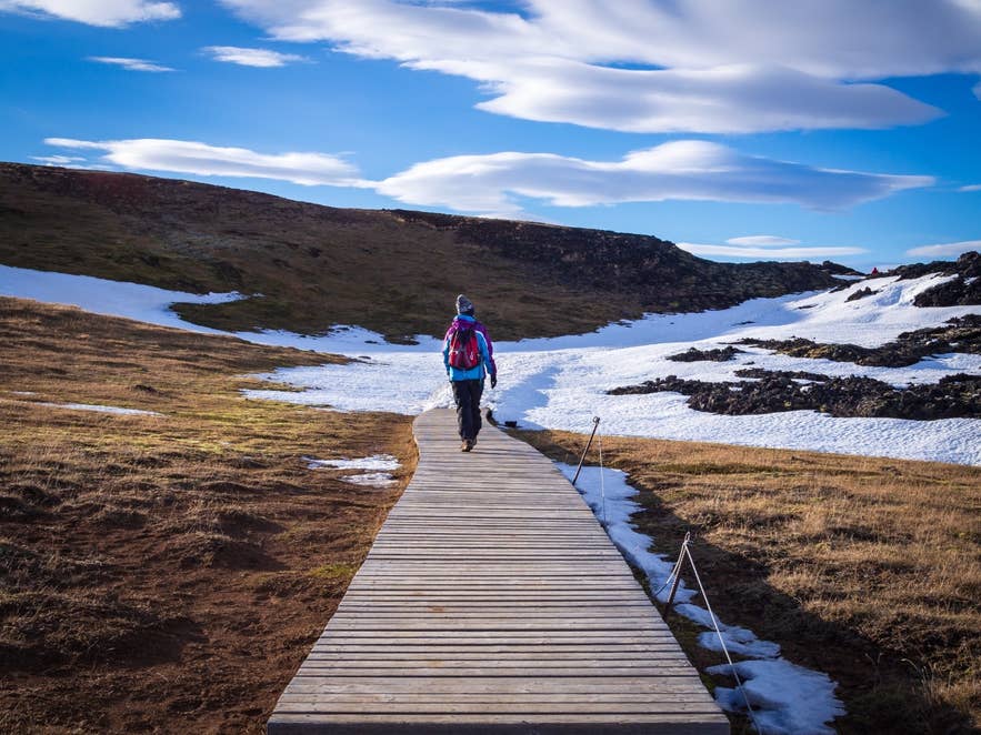 A hiker walks a wooden boardwalk through patchy snow on a spring day in North Iceland.