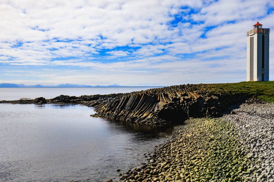 Lighthouse on grassy coast at Kálfshamarsvík, Iceland, with basalt rock columns, ocean, and distant mountains.