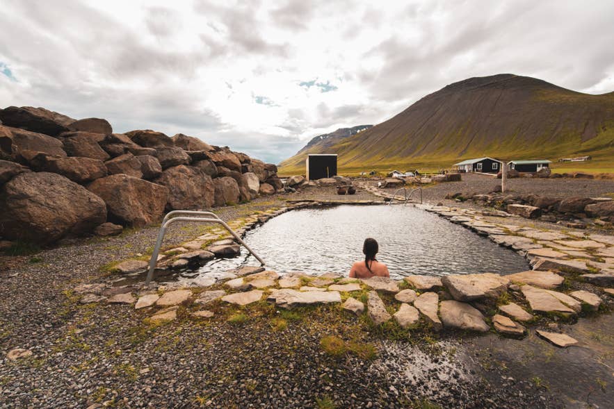 Grettislaug hot spring in Iceland, person bathing in stone pool with mountains and cloudy sky in background.
