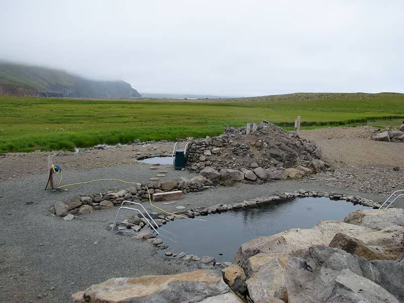 Stone-lined geothermal pool at Grettislaug, surrounded by open grassland, rocks, and misty coastal mountains.