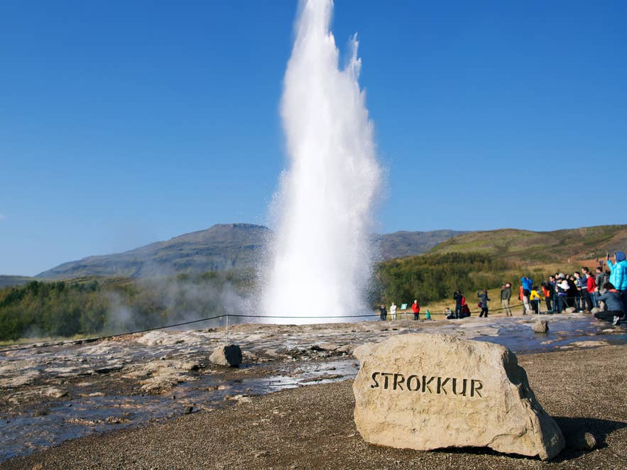 Strokkur Geyser erupts behind a stone marked with its name as visitors watch in Haukadalur, Iceland.