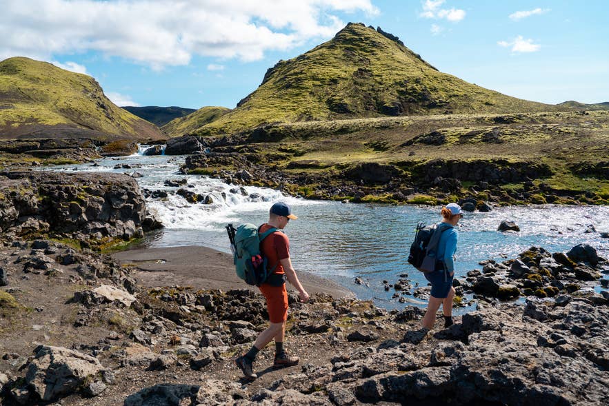 Two hikers with backpacks reaching a glacial river crossing beside rapids on the Laugavegur Trail