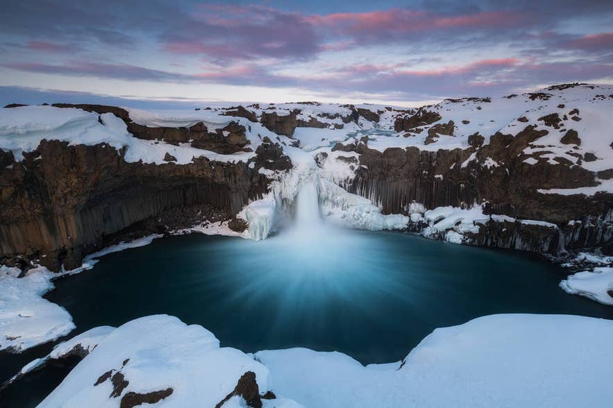 De waterval Aldeyjarfoss in IJsland tijdens de winter, omringd door ijzige basaltzuilen en een besneeuwd landschap.