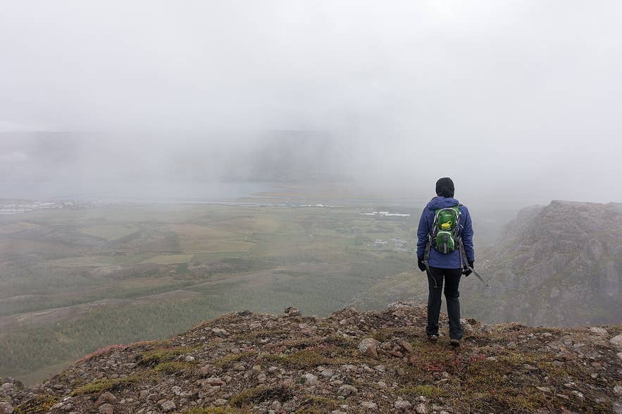Hiker on rocky cliffs above Akureyri, overlooking Naustaborgir hiking area with misty valley and fjord views.