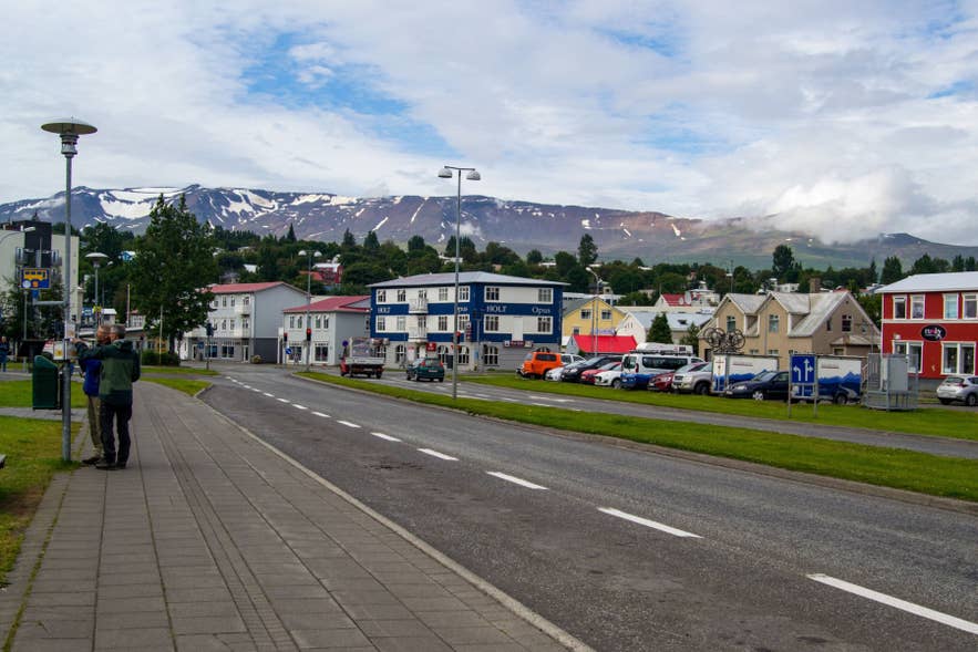 Colorful Akureyri street with HOLT Opus, parked cars, bus stop, and snowy mountains rising in the background.