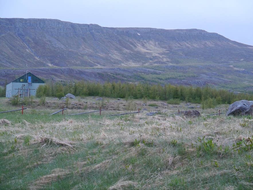 Small green hut in grassy Akureyri field with rocky mountain ridge rising under an overcast sky.