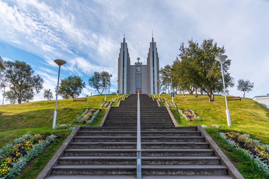 Staircase lined with flowers leading up to Akureyrarkirkja church towers under a partly cloudy sky.