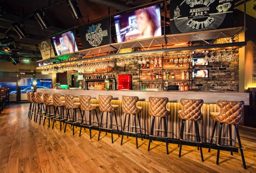 Interieur van The American Bar Reykjavik met leren barkrukken met knopen, live schermen en een volledig uitgeruste cocktailbar.