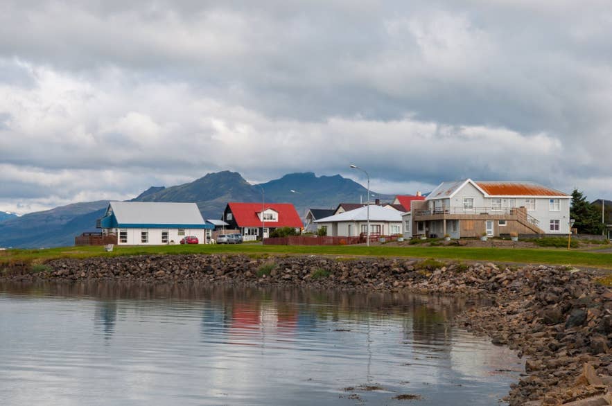 Colorful houses along the shoreline in Hofn with calm water and mountains in East Iceland under cloudy skies.