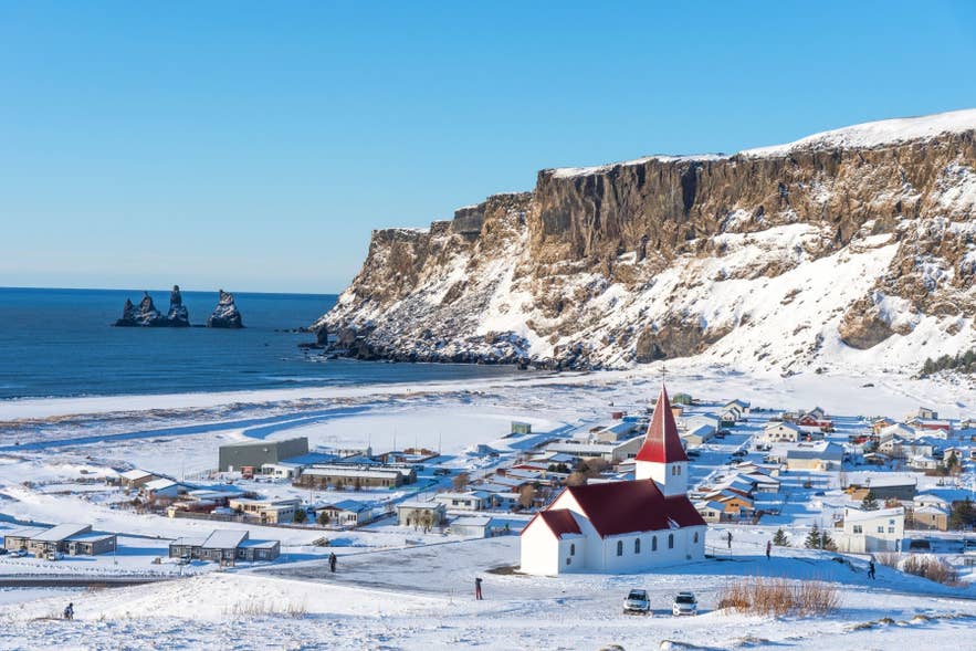 Die Kirche von Vik i Myrdal mit Blick auf verschneite Klippen, schwarze Sandküste und die Reynisdrangar-Felsnadeln in Südisland.