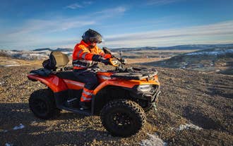 Person in high-visibility gear riding an ATV on a rocky hilltop with panoramic views of the Icelandic Highlands and snow-dusted mountains in the distance.