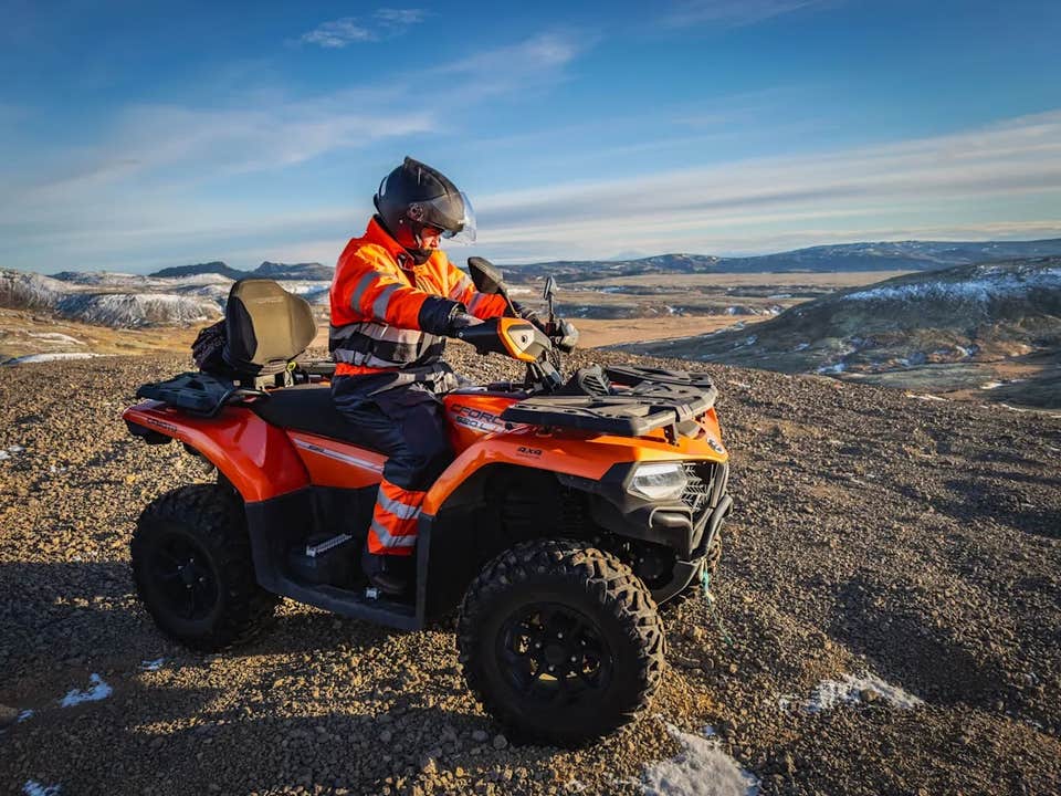 Person in high-visibility gear riding an ATV on a rocky hilltop with panoramic views of the Icelandic Highlands and snow-dusted mountains in the distance.