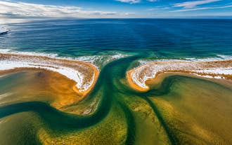 Aerial view of winding turquoise tidal channels flowing through Raudasandur beach in the Westfjords, with golden sand, patches of snow, and the Atlantic Ocean beyond.