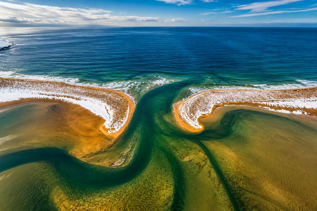 Aerial view of winding turquoise tidal channels flowing through Raudasandur beach in the Westfjords, with golden sand, patches of snow, and the Atlantic Ocean beyond.
