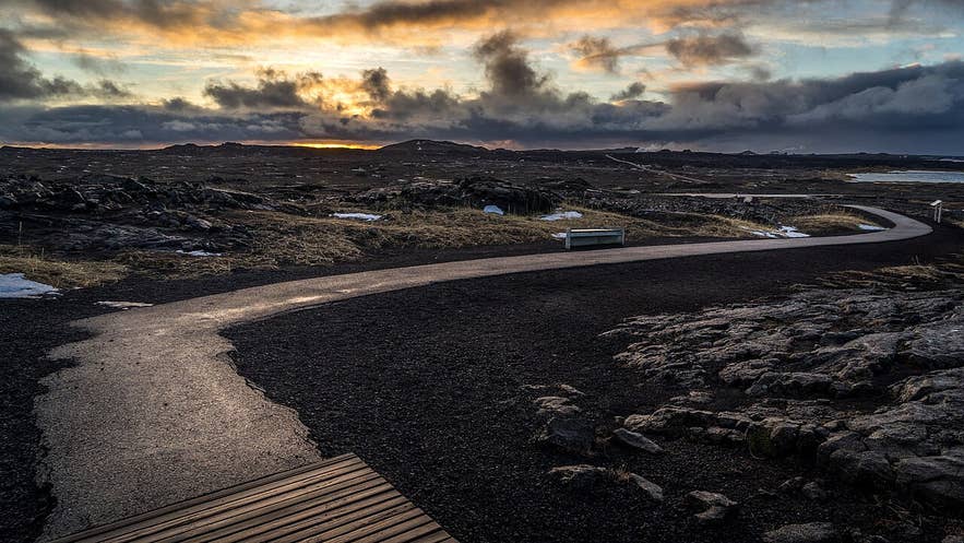 Winding path at Sandvík Beach, Reykjanes Peninsula, with volcanic terrain, bench, and golden sunset sky.