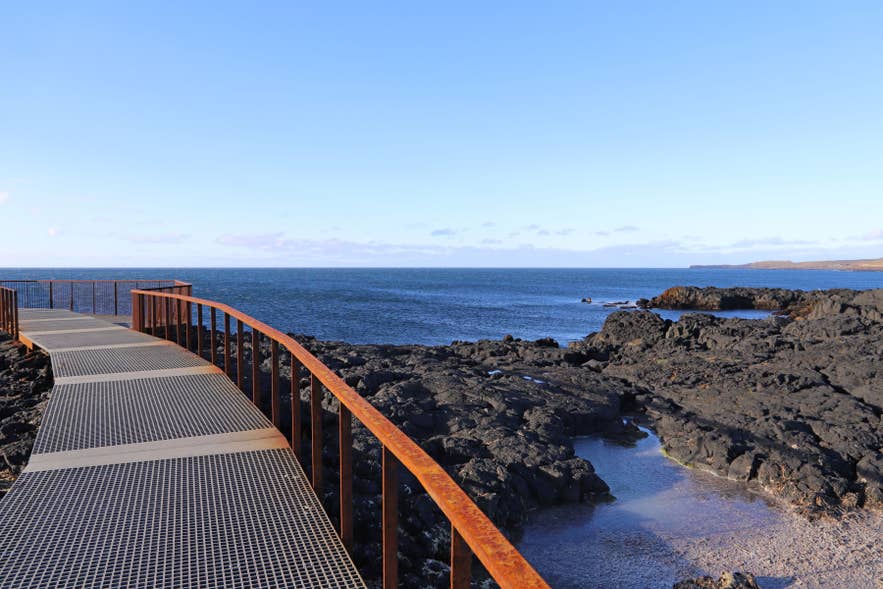 Rust‑colored walkway along volcanic shore, overlooking Brimketill lava rock pool on Reykjanes Peninsula