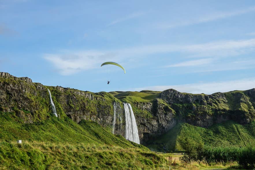 A paraglider soars above Seljalandsfoss waterfall, with green hills and cascading water along Iceland’s South Coast.