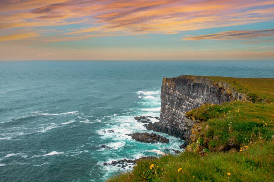 Latrabjarg sea cliffs rise above the Atlantic Ocean in the Westfjords, with waves crashing below beneath a colorful sunset sky.