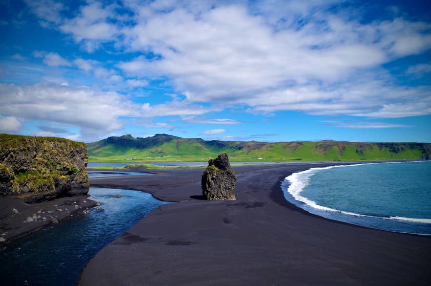 A basalt sea stack stands on a black sand beach near Dyrholaey, with waves, volcanic cliffs, and green mountains along Iceland’s South Coast.