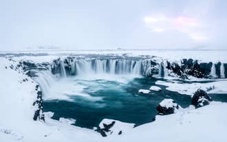 Aerial view of the partly frozen Godafoss Waterfall surrounded by snow in North Iceland.