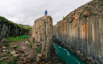 A traveller in a blue jacket stands atop the basalt columns overlooking Studlagil Canyon in East Iceland.