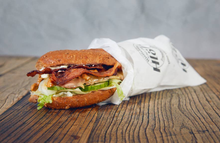 Warme sub van Hlollabatar met bacon, kip, sla, komkommer en saus op een geroosterd broodje, verpakt in bedrukt papier op een houten oppervlak.