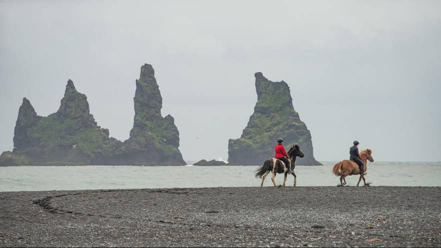 Horseback riders on Reynisfjara black sand beach, with the Reynisdrangar sea stacks rising from the ocean in the background.